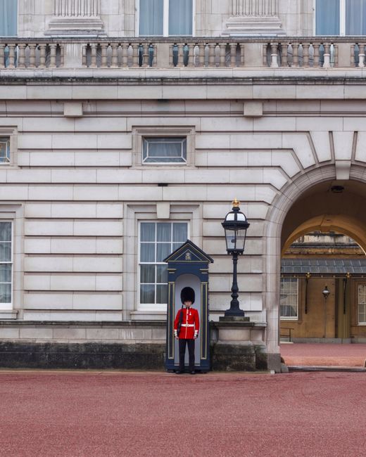A member of Westminster Removals staff dressed in a traditional red uniform with a tall black bearskin hat standing inside a blue wooden guardhouse positioned on a pavement in front of a historic ston